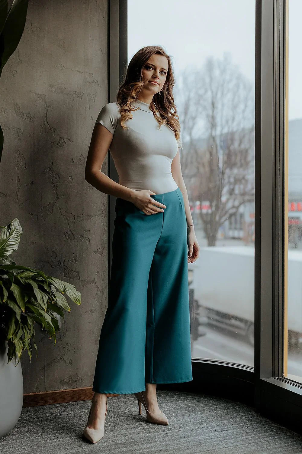 Woman in teal cropped wide-leg trousers and a fitted top by a window fashion image